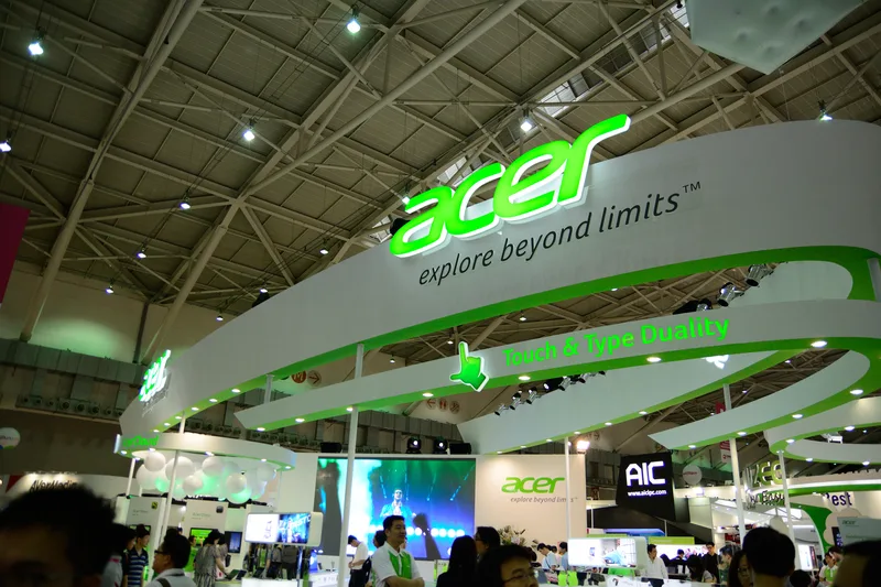 Suasana booth pameran komputer dan teknologi di aula konvensi dengan meja display laptop, lampu truss, dan videotron panggung